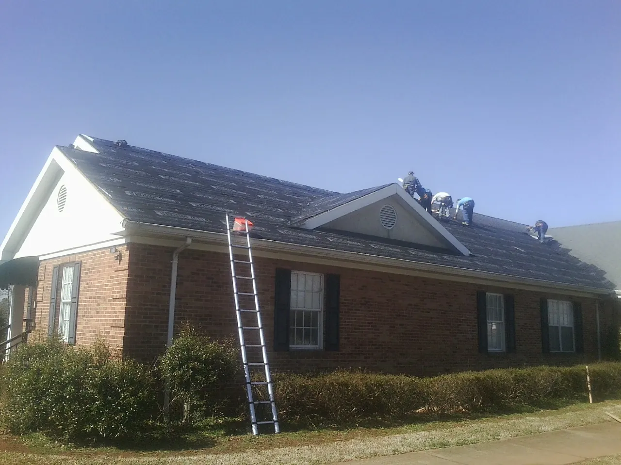 Crew tearing shingles off a brick ranch roof with a ladder set — Metal Roof Repair work by our crew in Gibbon Glade