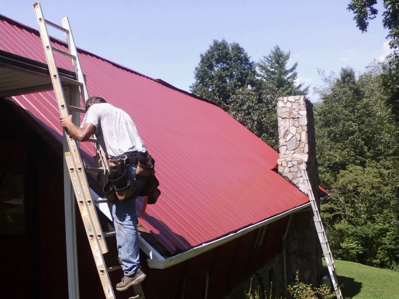 Roofer on a ladder beside a red metal roof and stone chimney — Soffit & Fascia Repair by Roofing Brothers in Gibbon Glade